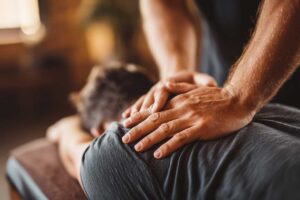 A chiropractor gently examines a patient’s back in a calm, sunlit treatment room.